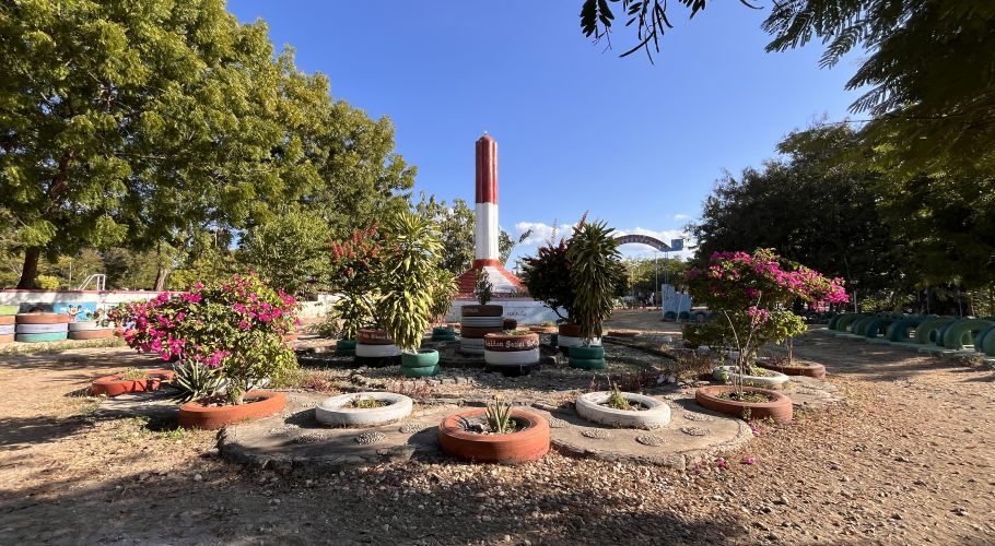 A red and white monument stands in a park surrounded by green trees, pink flowers, and tire planters under a clear blue sky.