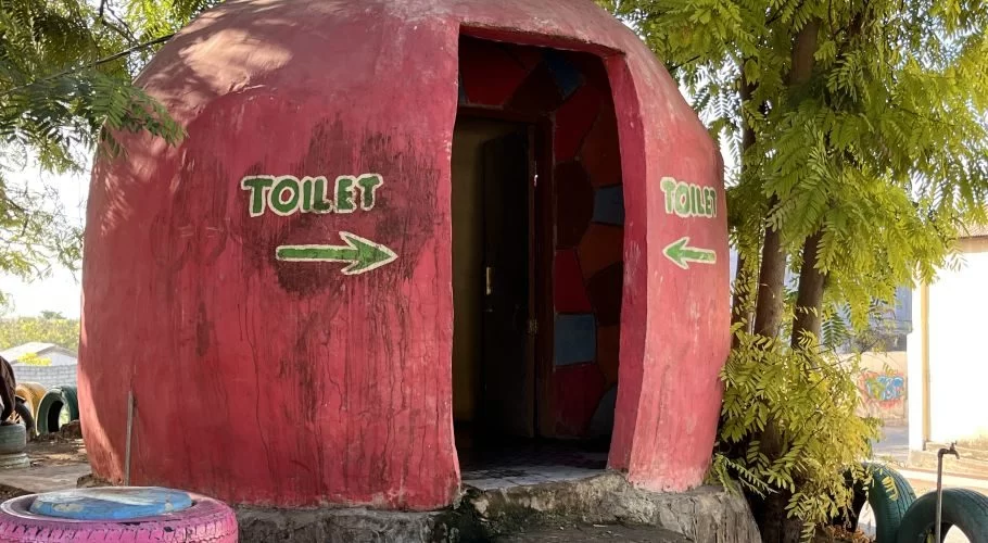 A red, dome-shaped public toilet building with green "TOILET" signs and arrows stands in an outdoor park setting. Its open doorway reveals colorful mosaic tiles, surrounded by green trees and repurposed tires.