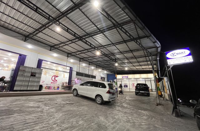 The brightly lit exterior of Cinta Karya Mart, a modern convenience store in Waingapu, Sumba, features a large parking area with a white car and a motorbike at night.
