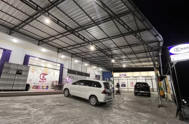 The brightly lit exterior of Cinta Karya Mart, a modern convenience store in Waingapu, Sumba, features a large parking area with a white car and a motorbike at night.