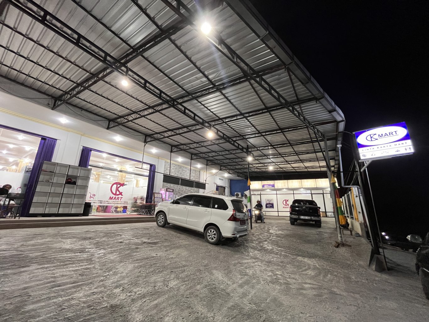 The brightly lit exterior of Cinta Karya Mart, a modern convenience store in Waingapu, Sumba, features a large parking area with a white car and a motorbike at night.