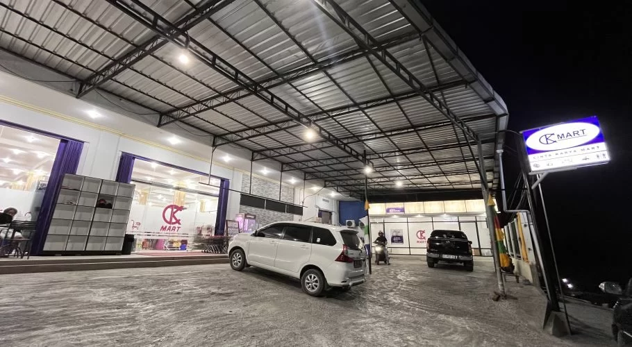 The brightly lit exterior of Cinta Karya Mart, a modern convenience store in Waingapu, Sumba, features a large parking area with a white car and a motorbike at night.