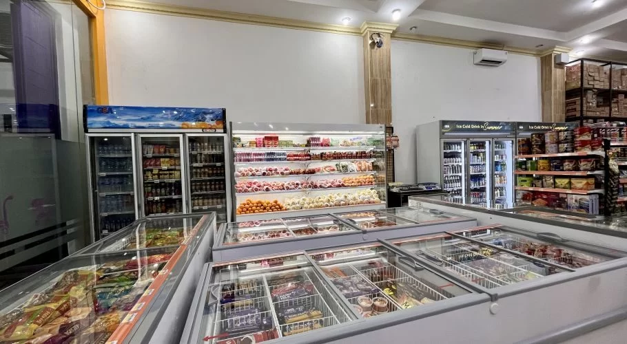 Brightly lit interior of Cinta Karya Mart, a grocery store in Waingapu, showing rows of refrigerators with drinks, fresh produce, and freezers with packaged goods.