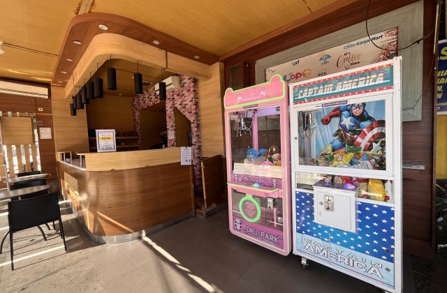 Two colorful arcade claw machines, one pink with plush toys and one Captain America-themed, stand next to a wooden service counter in a bright play area.