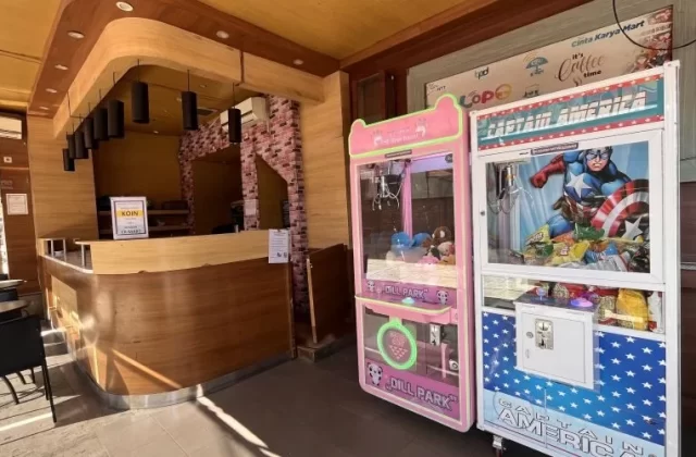 Two colorful arcade claw machines, one pink with plush toys and one Captain America-themed, stand next to a wooden service counter in a bright play area.