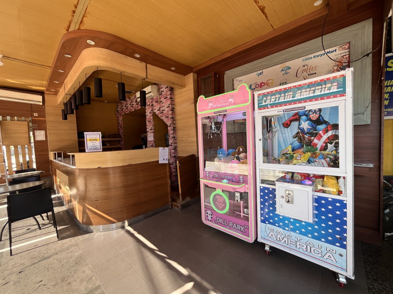 Two colorful arcade claw machines, one pink with plush toys and one Captain America-themed, stand next to a wooden service counter in a bright play area.