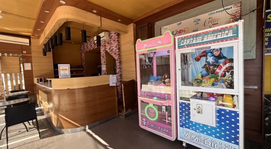Two colorful arcade claw machines, one pink with plush toys and one Captain America-themed, stand next to a wooden service counter in a bright play area.