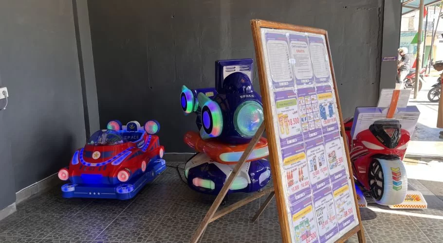 Three colorful coin-operated kiddie rides, including a red car and blue spaceship, stand on a tiled floor next to a purple price board at an outdoor play area.