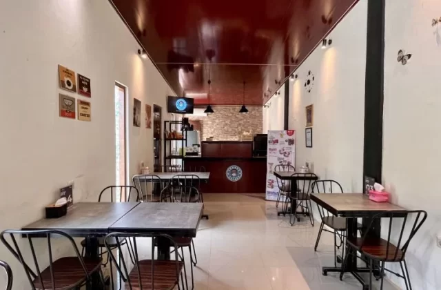 A long, narrow cafe interior features white walls, a dark red ceiling, and several black tables with matching chairs, leading to a dark wooden counter at the back.