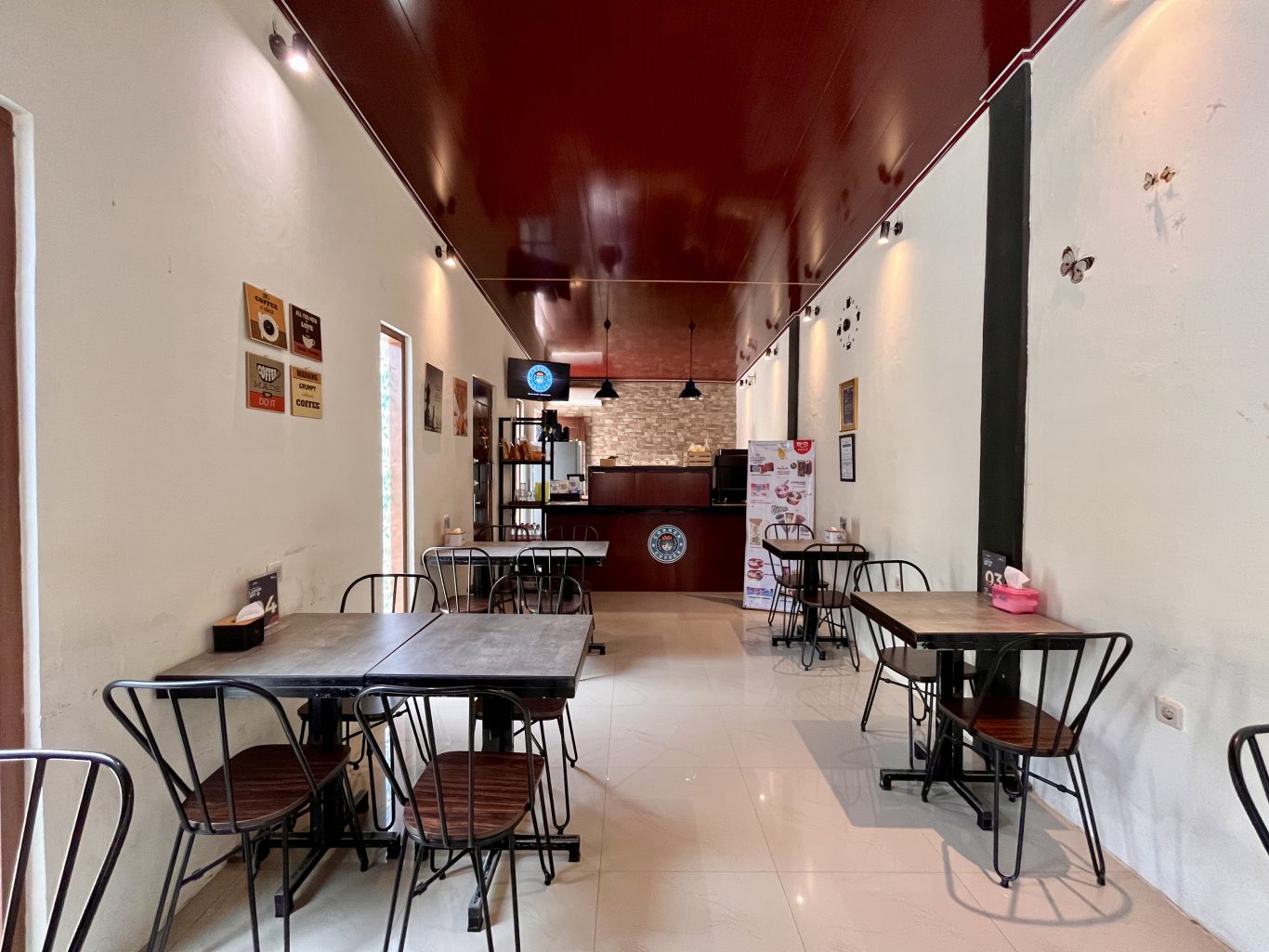 A long, narrow cafe interior features white walls, a dark red ceiling, and several black tables with matching chairs, leading to a dark wooden counter at the back.