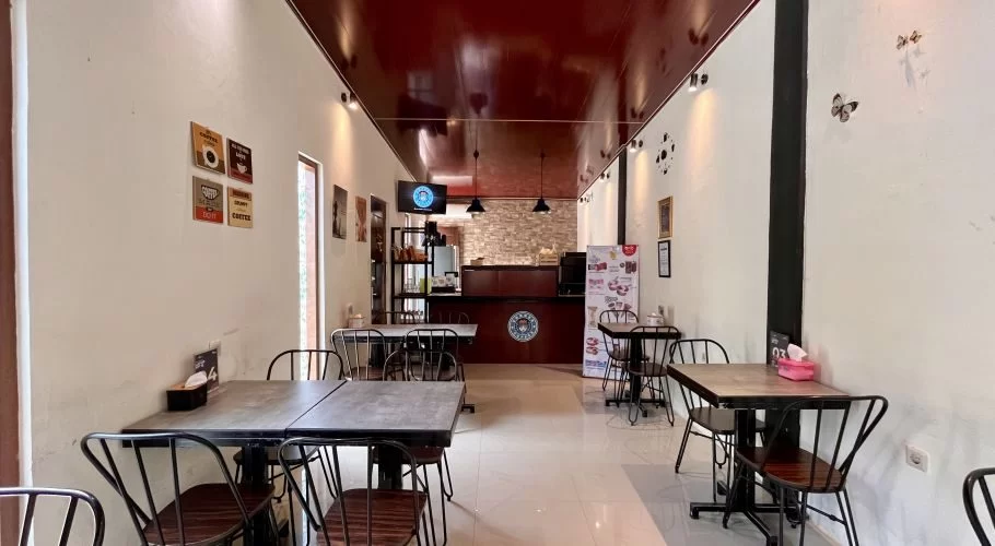 A long, narrow cafe interior features white walls, a dark red ceiling, and several black tables with matching chairs, leading to a dark wooden counter at the back.