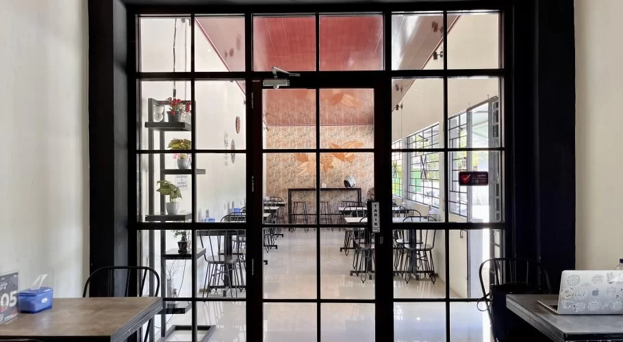 A view from inside a cafe, looking through black-framed glass doors into a brightly lit seating area with black tables, chairs, and a golden fish mural on a patterned wall.
