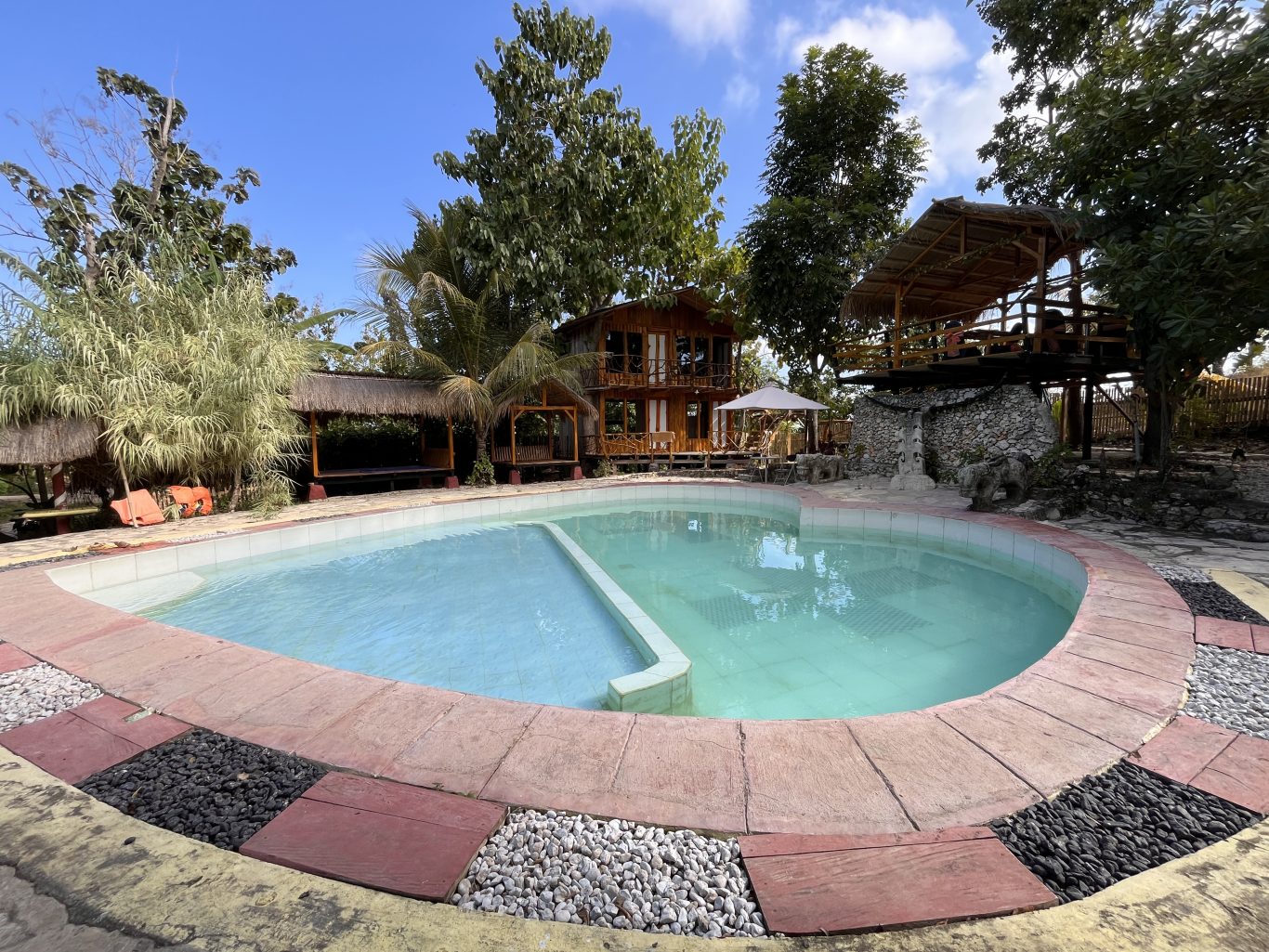 A light blue swimming pool with reddish-brown coping is surrounded by lush green trees, a two-story wooden building, and a thatched-roof treehouse under a clear blue sky.