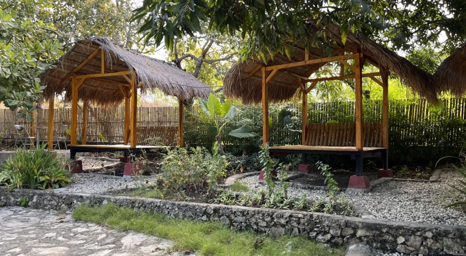 Visible are two traditional Sumbanese gazebos with thatched roofs and wooden frames, surrounded by lush green foliage and gravel paths under a bright sky.