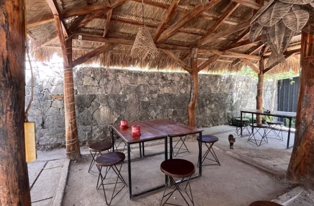 An open-air restaurant dining area features a thatched roof, thick wooden posts, stone walls, and dark wooden tables with stools. Two red candle holders are on a table, and a small cat walks in the background.