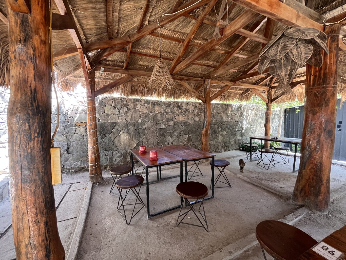 An open-air restaurant dining area features a thatched roof, thick wooden posts, stone walls, and dark wooden tables with stools. Two red candle holders are on a table, and a small cat walks in the background.