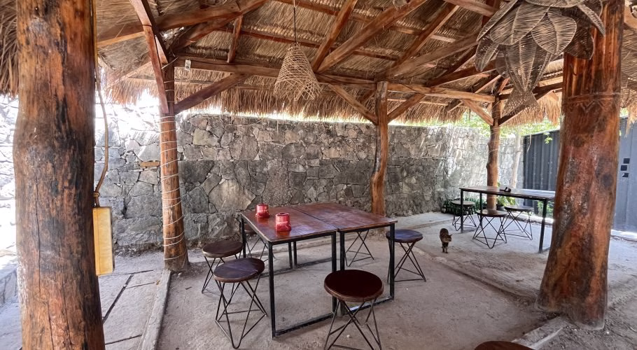 An open-air restaurant dining area features a thatched roof, thick wooden posts, stone walls, and dark wooden tables with stools. Two red candle holders are on a table, and a small cat walks in the background.