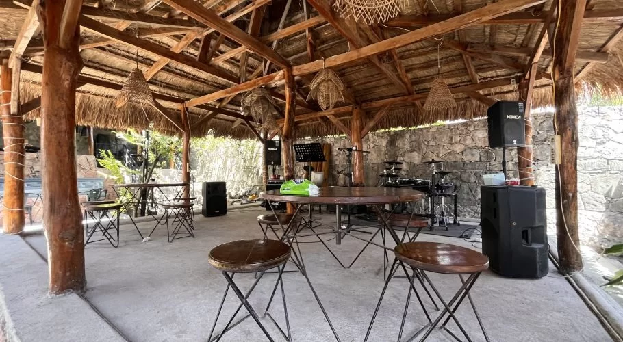 An open-air restaurant dining area in warm tones features a thatched roof, wooden posts, small tables, stools, and a stage with a drum set and speakers.