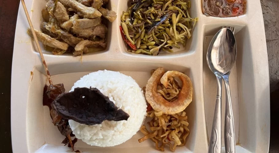 A compartmentalized white tray holds a meal with sliced pork, green vegetables, white rice, crispy pork skin, fried noodles, a meat skewer, and a small dish of chili sauce, with a spoon and fork.