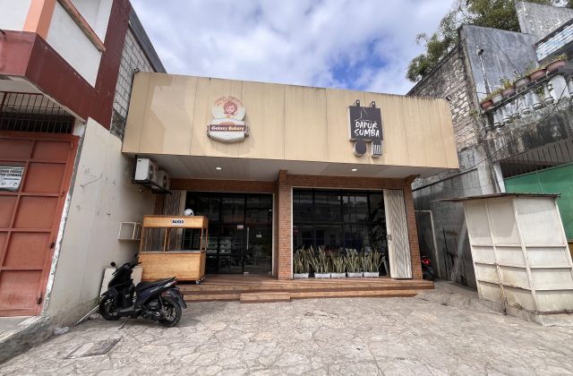 Exterior of Dapur Sumba restaurant in Waikabukak, Sumba, showing a light-colored building with glass windows and a bakso cart.