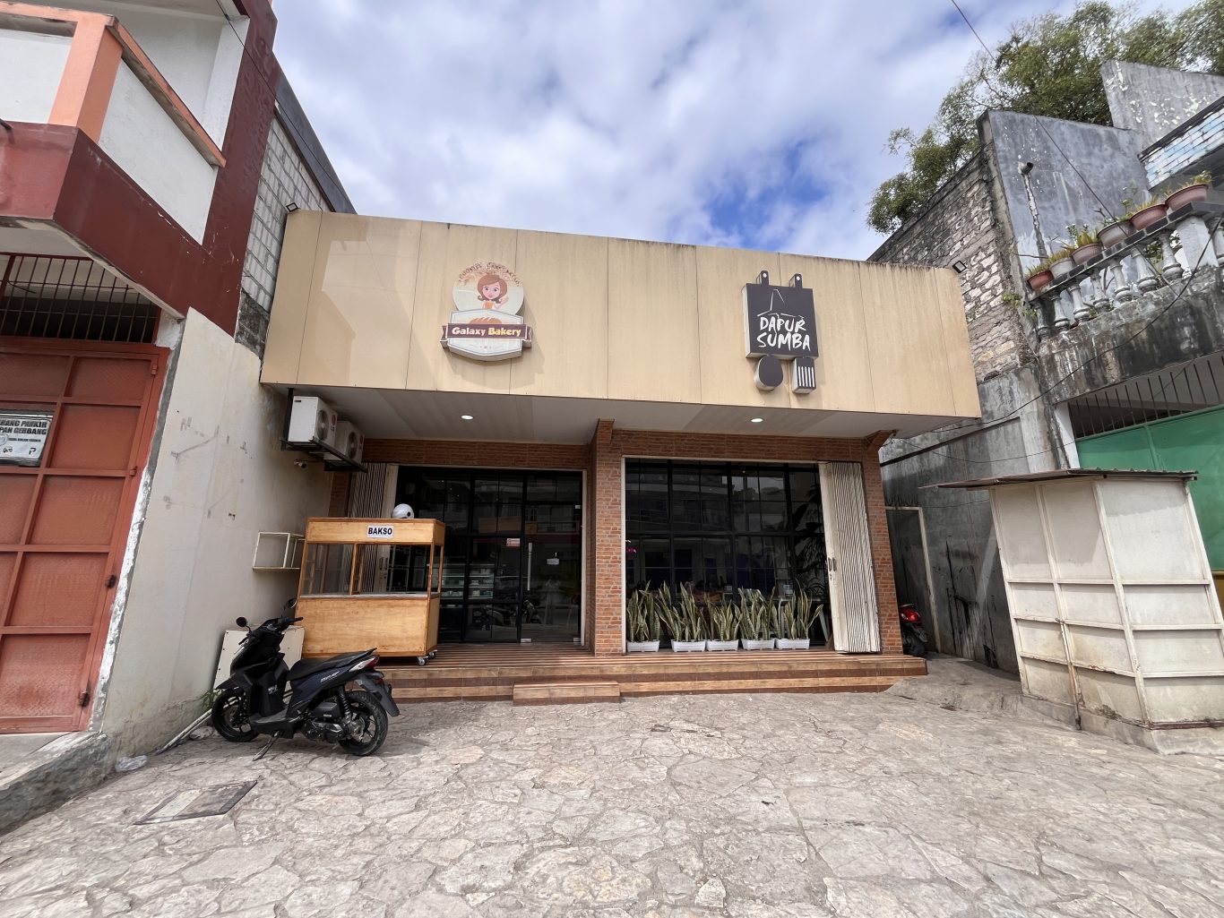 Exterior of Dapur Sumba restaurant in Waikabukak, Sumba, showing a light-colored building with glass windows and a bakso cart.