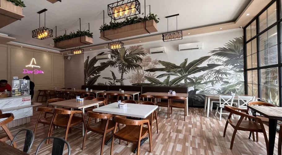 The spacious interior of Dapur Sumba restaurant features many wooden tables and chairs, a tropical leaf mural, hanging plants, and a pink neon sign.