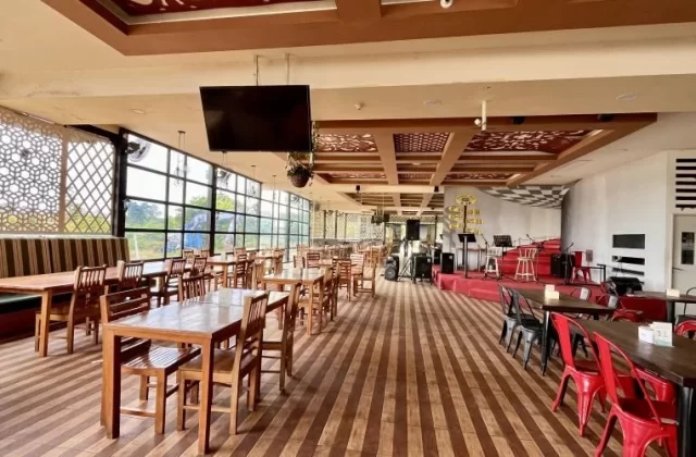 A spacious restaurant interior features numerous wooden tables and chairs, a stage with musical equipment, and large windows with decorative screens.