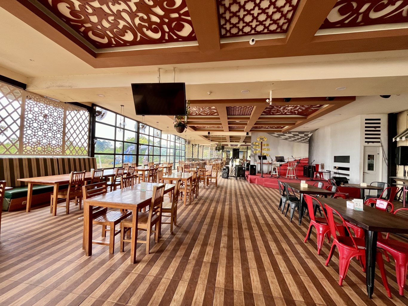 A spacious restaurant interior features numerous wooden tables and chairs, a stage with musical equipment, and large windows with decorative screens.