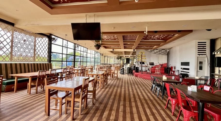 A spacious restaurant interior features numerous wooden tables and chairs, a stage with musical equipment, and large windows with decorative screens.