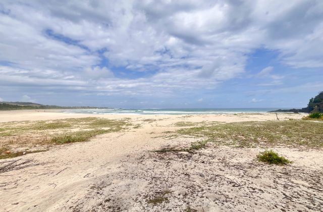 A wide view of Dasang Beach shows white sand with sparse green vegetation, gentle blue ocean waves, a dark rocky headland, and a partly cloudy sky.