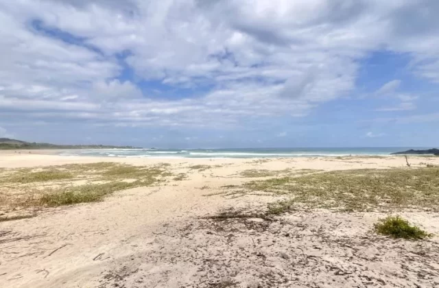 A wide view of Dasang Beach shows white sand with sparse green vegetation, gentle blue ocean waves, a dark rocky headland, and a partly cloudy sky.
