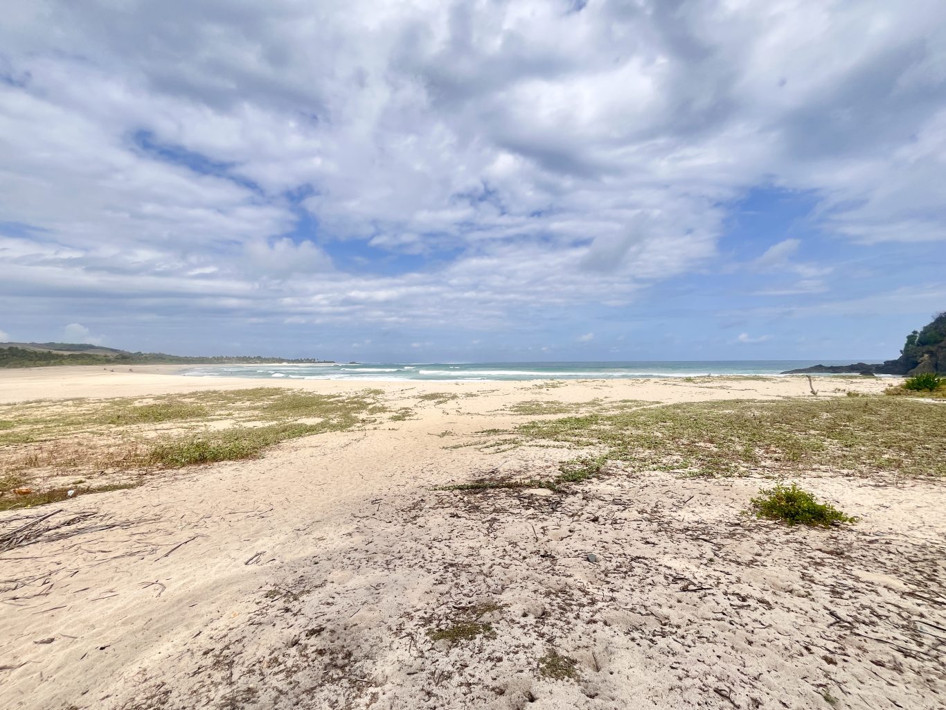 A wide view of Dasang Beach shows white sand with sparse green vegetation, gentle blue ocean waves, a dark rocky headland, and a partly cloudy sky.