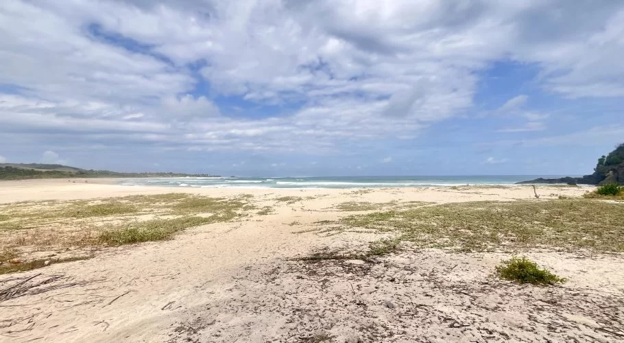 A wide view of Dasang Beach shows white sand with sparse green vegetation, gentle blue ocean waves, a dark rocky headland, and a partly cloudy sky.