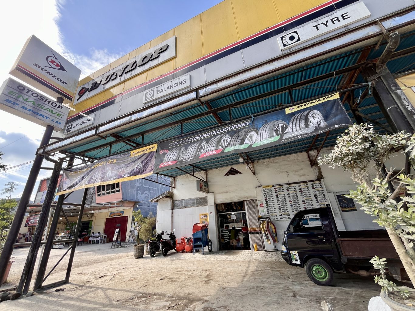 The yellow and white exterior of Delta Motor, a car mechanic shop in Waikabubak, features Dunlop signs, tire banners, and parked vehicles under a bright blue sky.