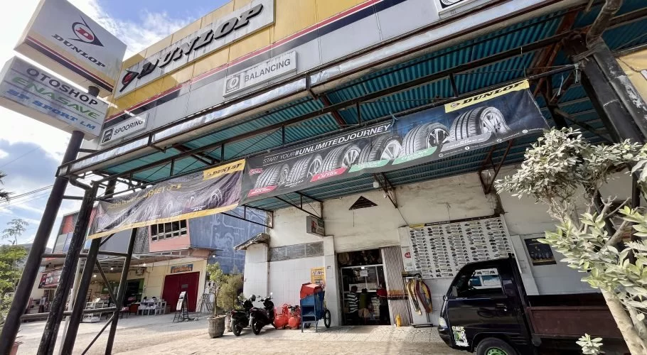 The yellow and white exterior of Delta Motor, a car mechanic shop in Waikabubak, features Dunlop signs, tire banners, and parked vehicles under a bright blue sky.