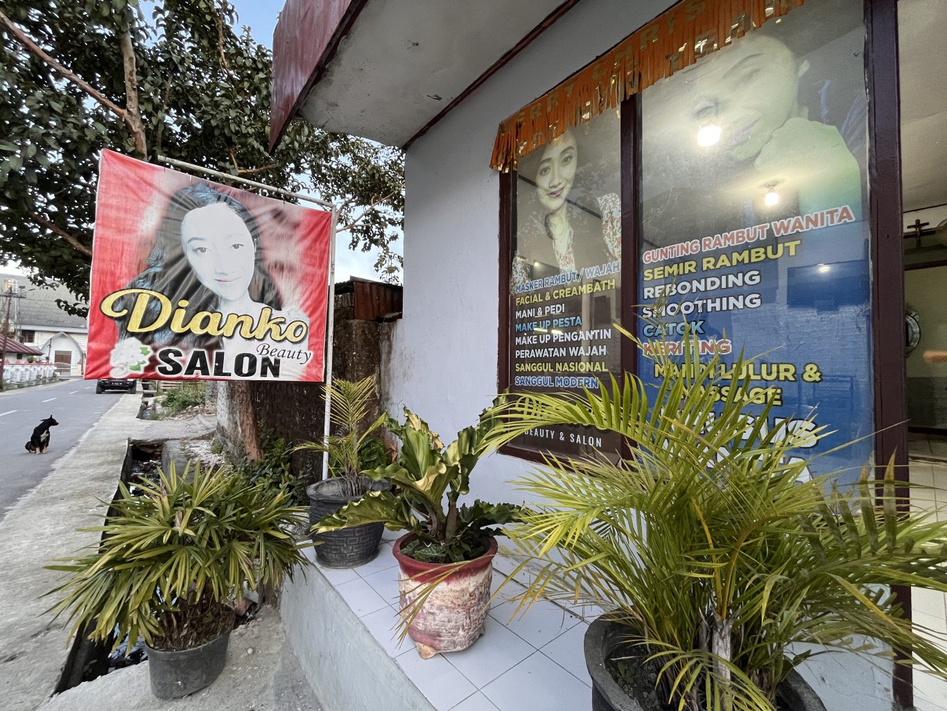 The exterior of Dianko Salon in Waikabubak, West Sumba, features a red banner and a window listing beauty services, with green potted plants and a street on the left.