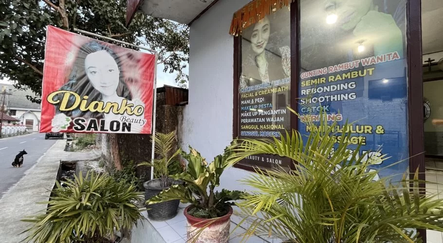 The exterior of Dianko Salon in Waikabubak, West Sumba, features a red banner and a window listing beauty services, with green potted plants and a street on the left.