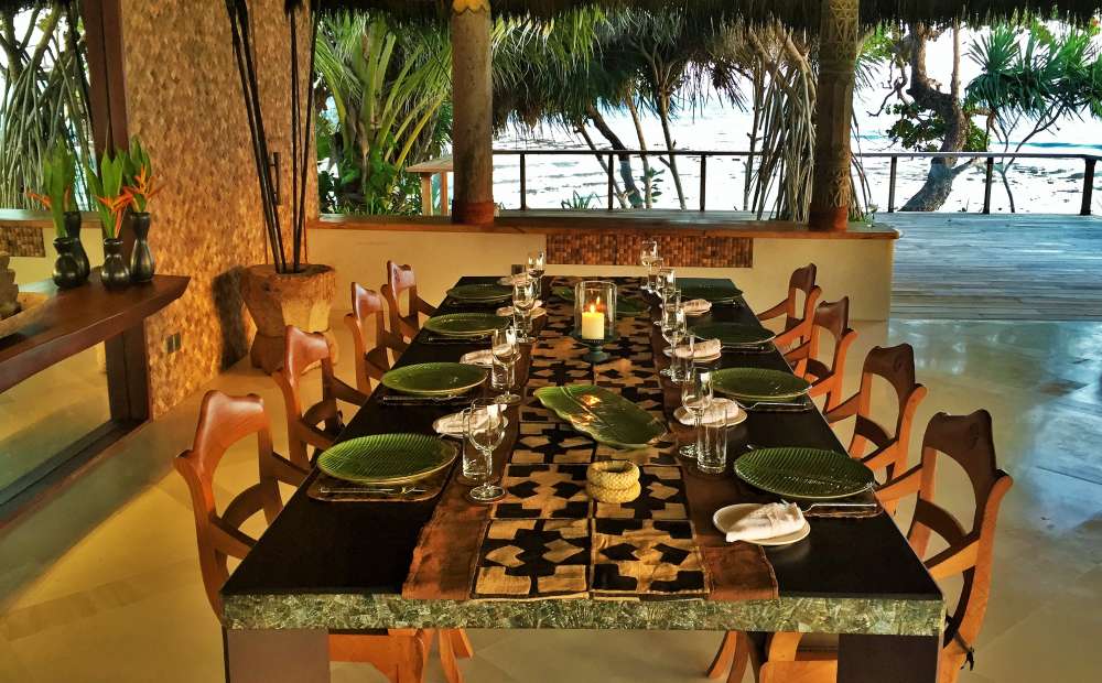 A long, dark dining table set with green plates and glasses sits in an open-air, thatched-roof dining area overlooking the ocean through tropical trees.
