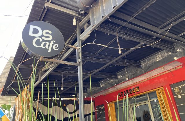 A black circular sign with 'DS Cafe' in white letters hangs from a metal frame above a red building wall and green plants, under a dark corrugated roof.