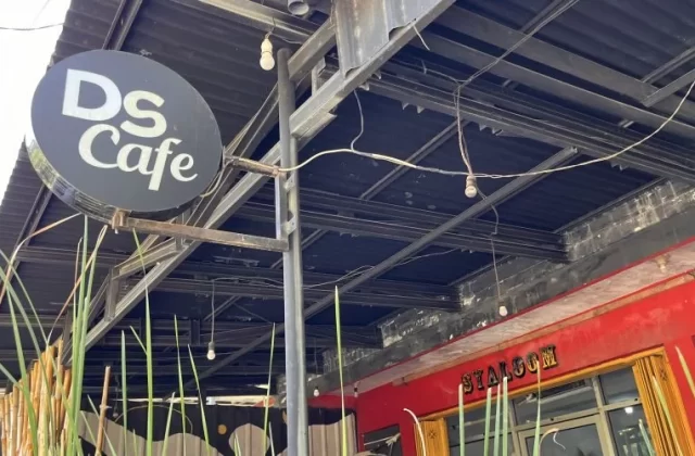 A black circular sign with 'DS Cafe' in white letters hangs from a metal frame above a red building wall and green plants, under a dark corrugated roof.