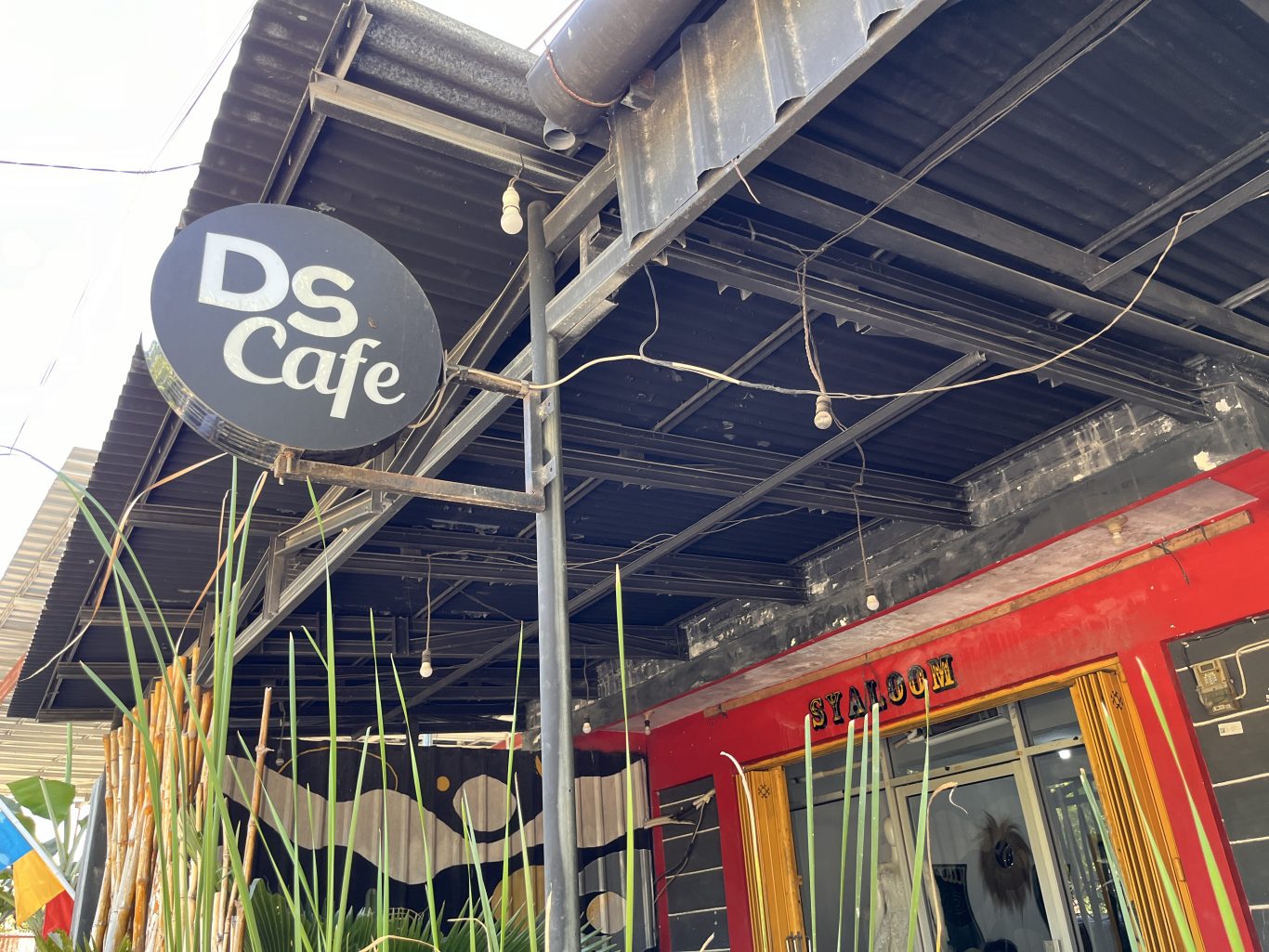 A black circular sign with 'DS Cafe' in white letters hangs from a metal frame above a red building wall and green plants, under a dark corrugated roof.
