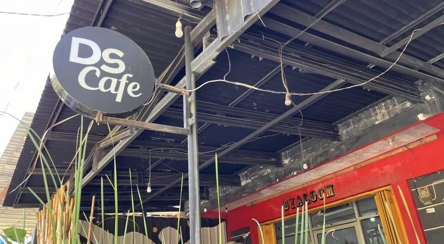 A black circular sign with 'DS Cafe' in white letters hangs from a metal frame above a red building wall and green plants, under a dark corrugated roof.