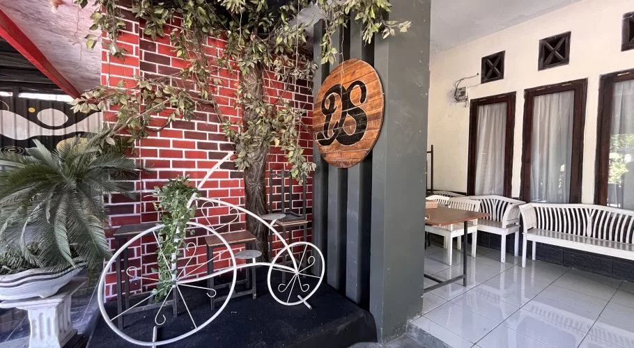 A cafe entrance features a red brick wall adorned with artificial vines, a white decorative bicycle, and a "DS" logo sign, with outdoor seating visible to the right.
