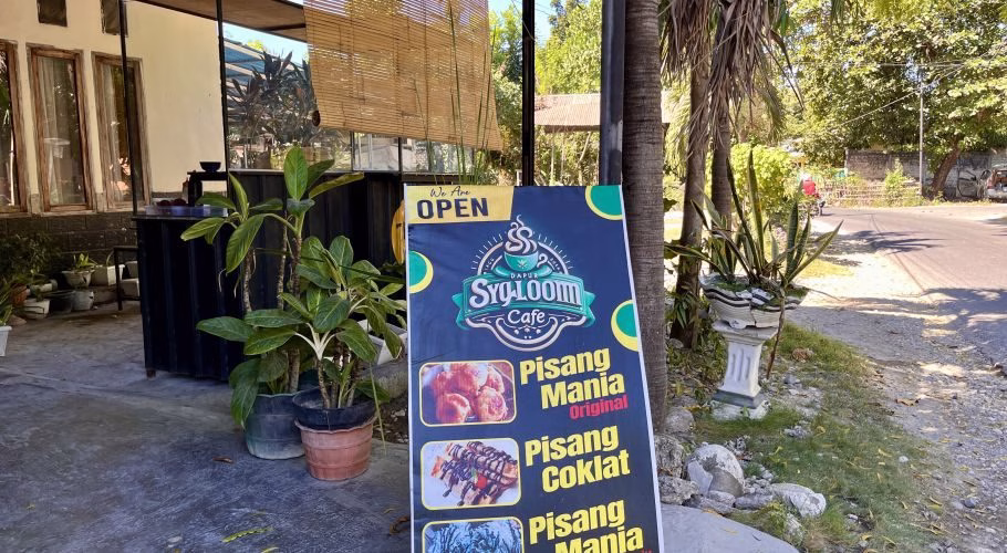 A blue and black 'Dapur Sygloom Cafe' sign, advertising banana snacks, stands outside a building with potted plants on a sunny day in Sumba.