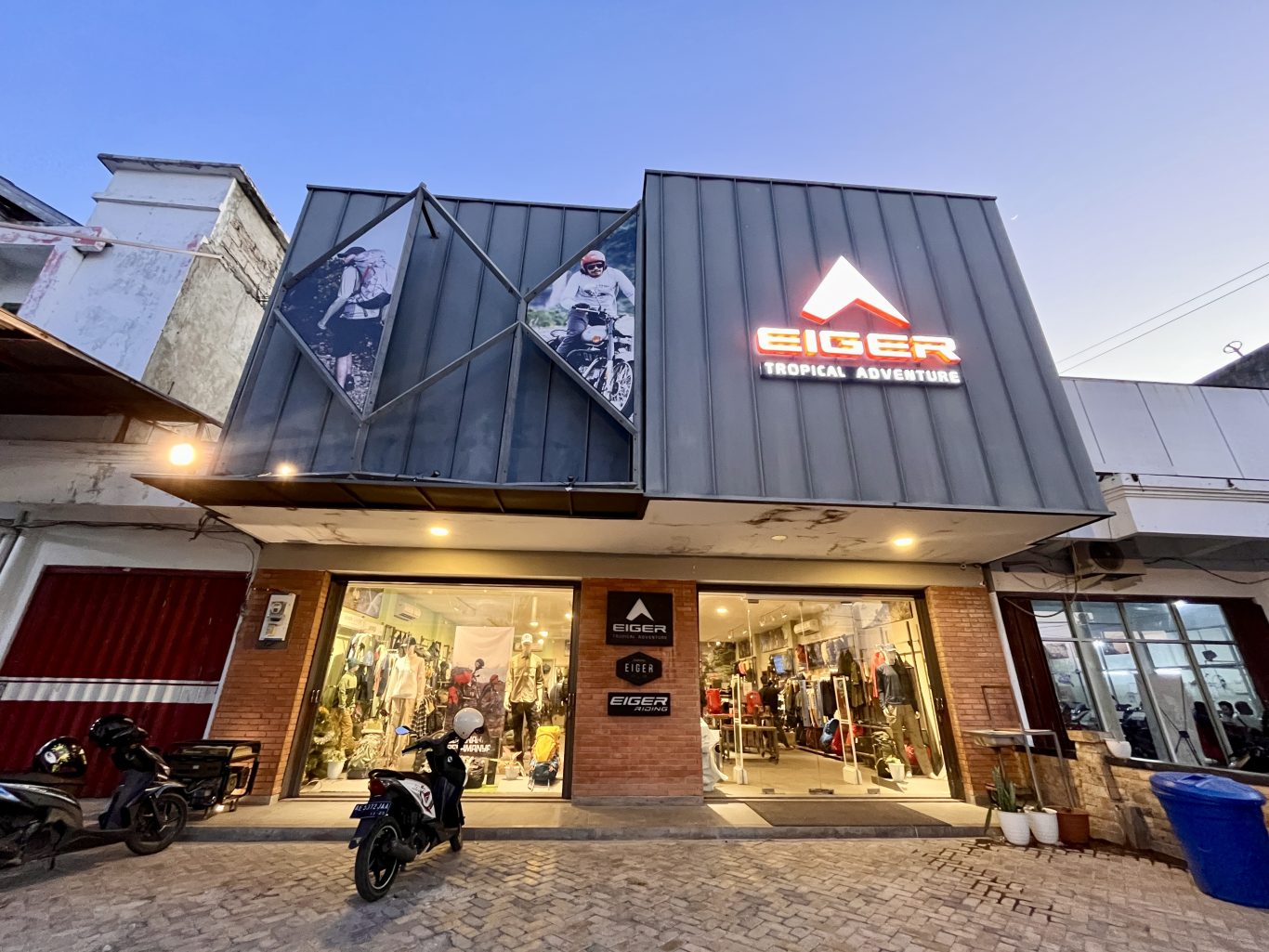 The Eiger outdoor gear store in Waingapu features a modern grey facade with a glowing orange logo, large display windows, and images of adventurers.