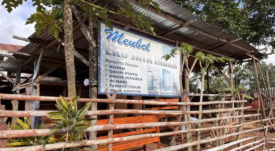 The exterior of Eko Jaya Indah Carpenter workshop in West Sumba features a blue and white sign, a corrugated metal roof, and a rustic wooden fence with green plants.