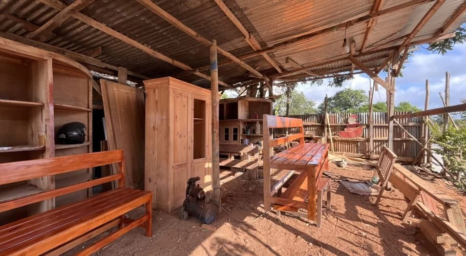 A rustic carpentry workshop in Sumba displays various unfinished wooden furniture, including benches, cabinets, and shelves, under a corrugated metal roof.