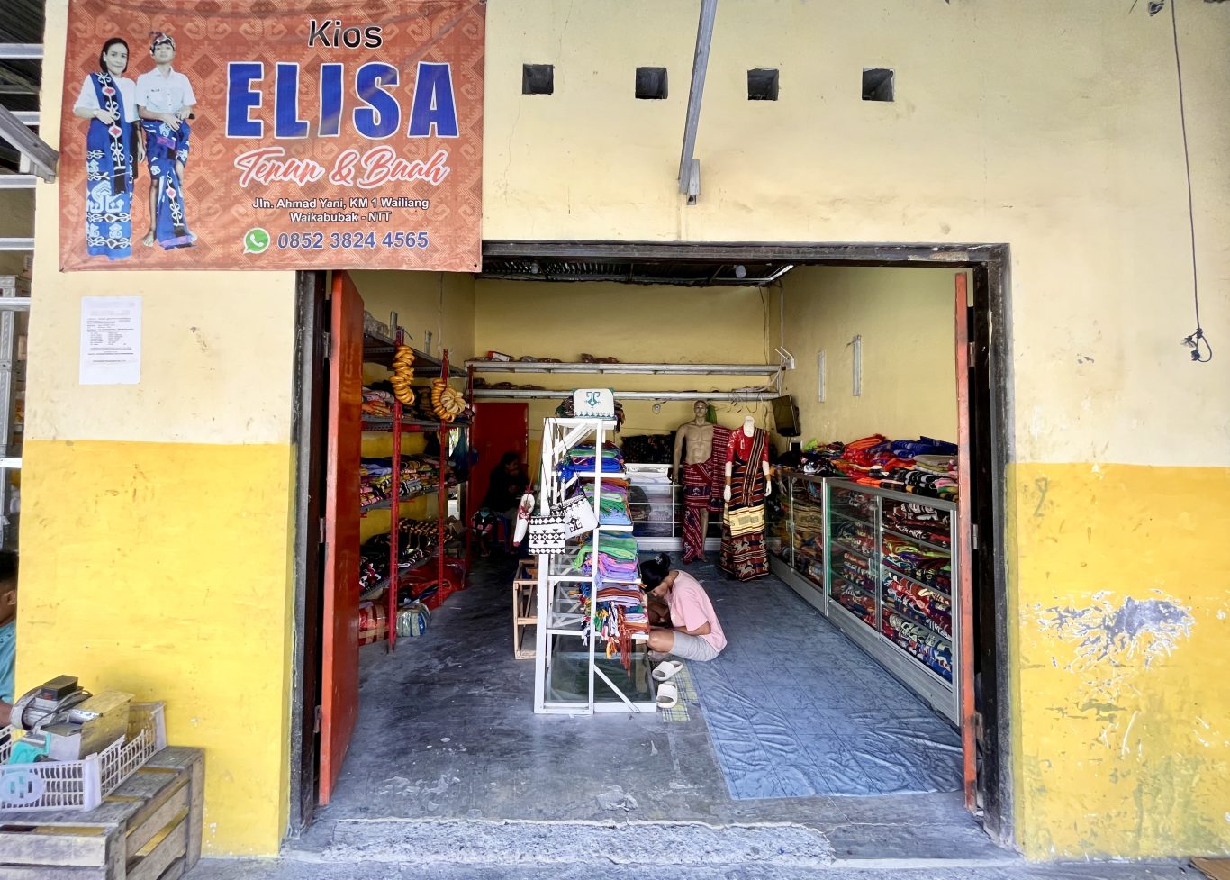 The entrance to Elisa Store reveals a person kneeling amidst vibrant Ikat Sumba textiles displayed on shelves and mannequins, under a banner.