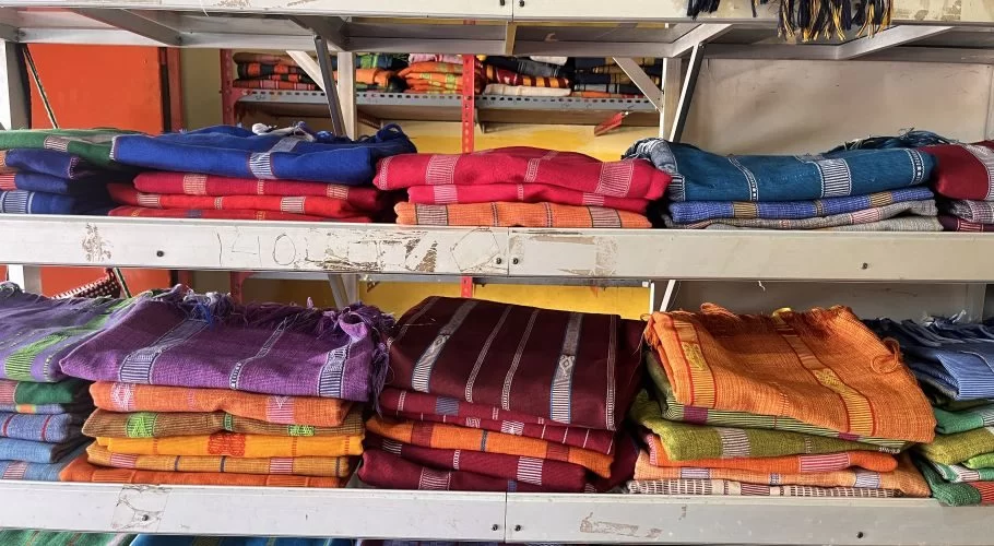 Stacks of vibrant blue, red, purple, orange, and green Ikat Sumba textiles with intricate patterns are neatly displayed on metal shelves in a store.
