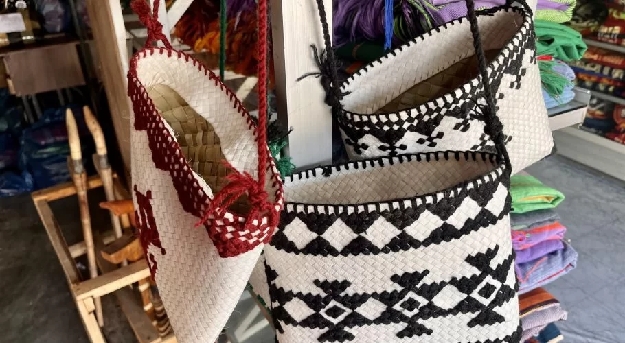 Several Sumba hand-woven bags with intricate white, black, and red patterns hang in a store, with colorful textiles behind.